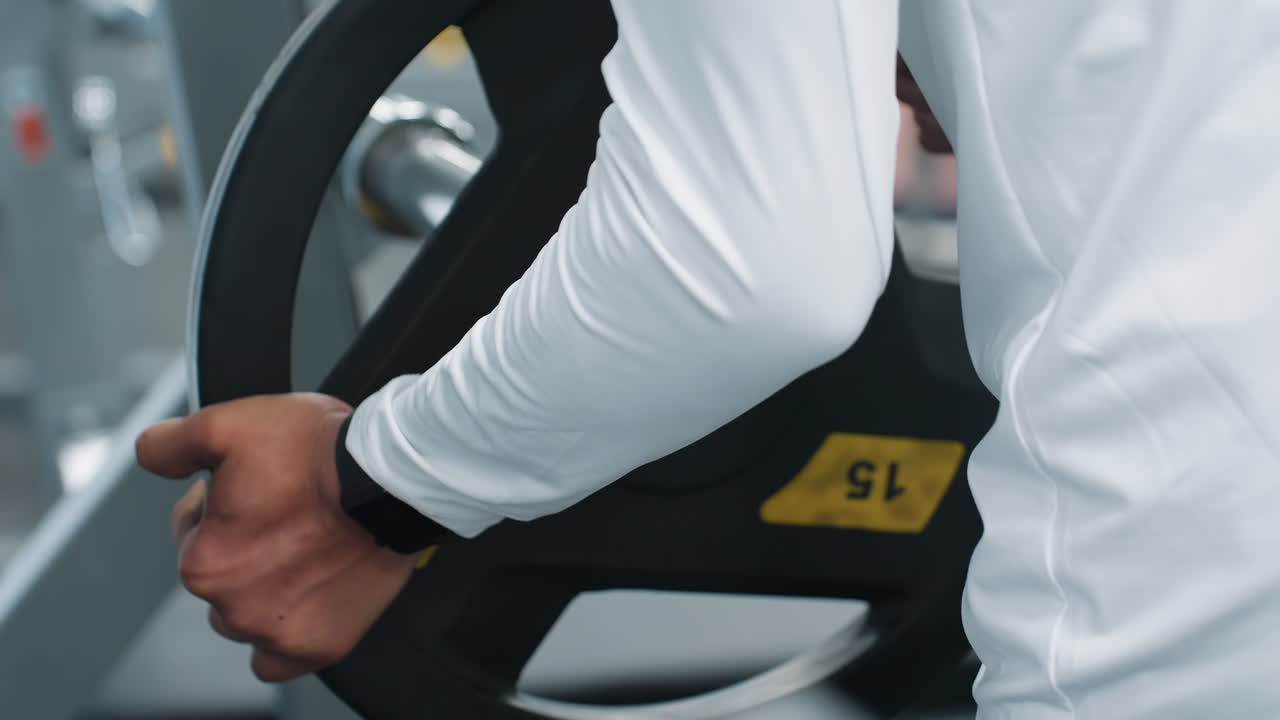 Rear close up of gym attendant returning weight plate into iron barbell rack on rubber flooring, showcasing hands guiding heavy plate into position during post workout equipment cleanup