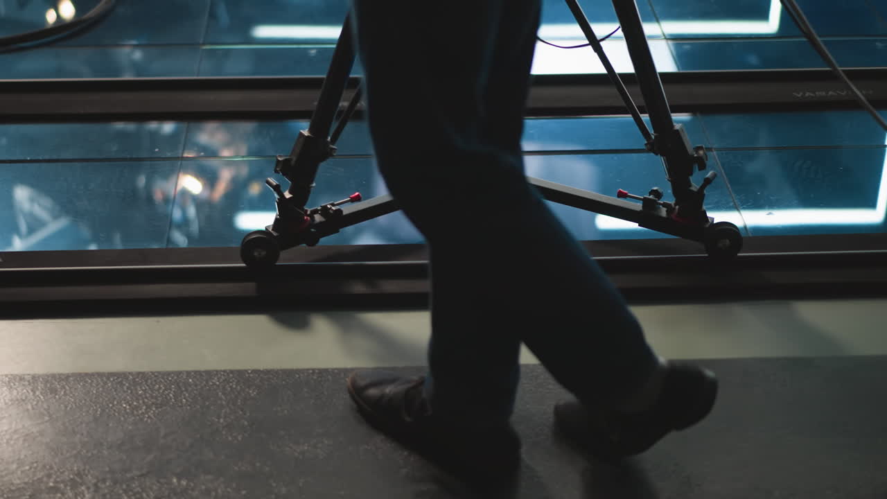 Close-up of camera dolly on track with operator s leg in motion, studio floor visible, broadcasting equipment in background, reflections on glass, lights shining through