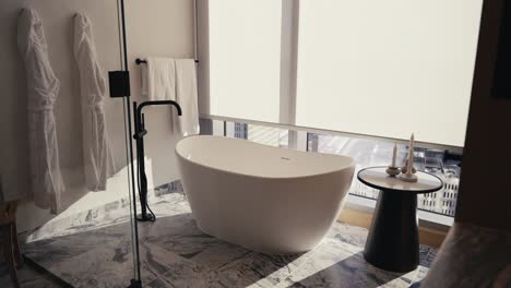 shades-rising-inside-of-a-luxury-bathroom-revealing-sunlight-to-showcase-a-free-standing-white-tub-with-a-side-table-along-with-towels-and-robes-hanging