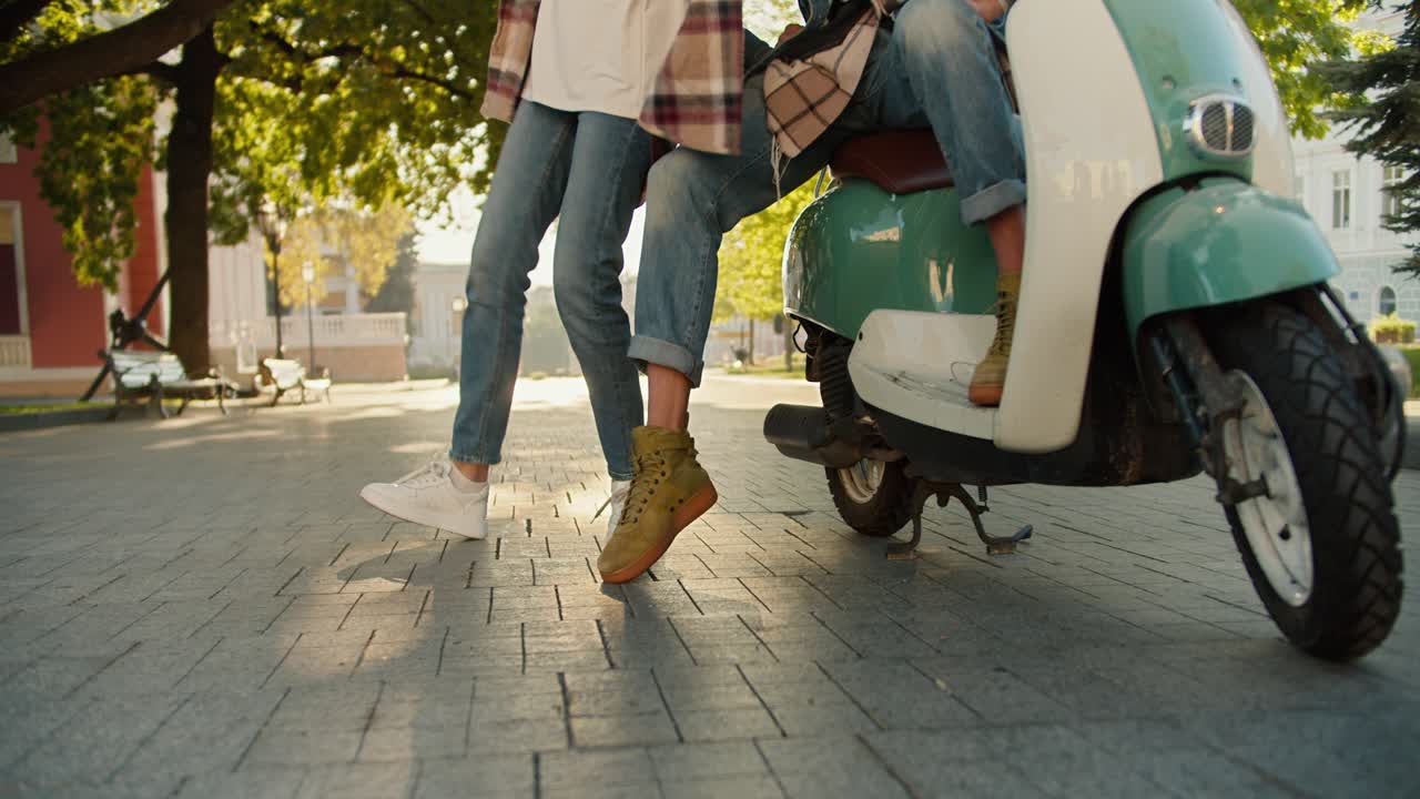 Close-up shot of a guy in jeans sitting on a green moped and his girlfriend in a checkered shirt, jeans and white sneakers dancing near him in a beautiful summer city park