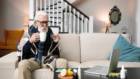 sick senior man sitting in room with cup of hot drink and videochatting on laptop