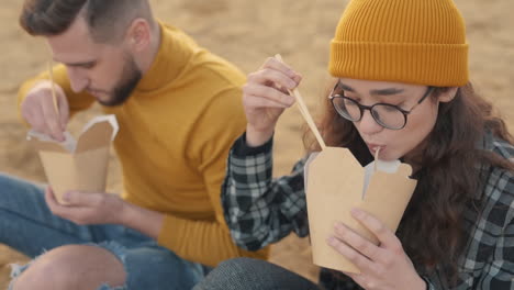 Young-Female-And-Male-Friends-Eating-Take-Away-Food-Sitting-On-The-Beach-1