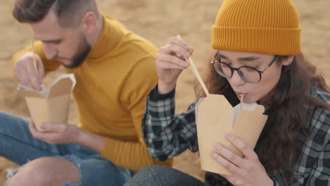 young female and male friends eating take away food sitting on the beach 1