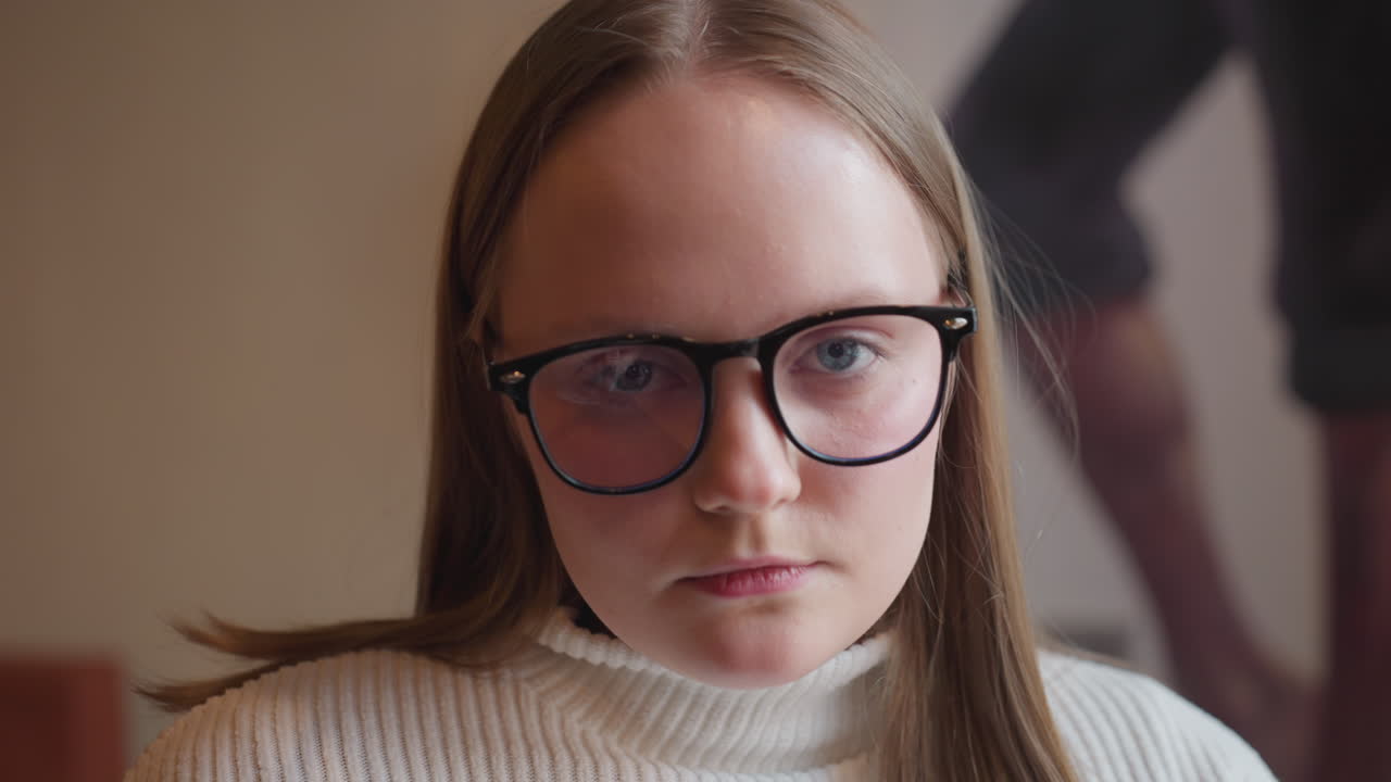 young woman in glasses wearing white ribbed sweater appears deep in thought as she looks down, warm decorative wall light softly illuminates background