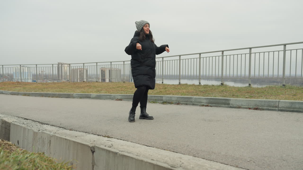Wanita menari di dekat pagar pembatas tepi air, mengenakan jaket puffer hitam dan kupluk rajut, berlatih gerakan gaya bebas di trotoar di bawah langit mendung, dengan latar belakang cakrawala kota, menampilkan gerakan ekspresif yang energik.