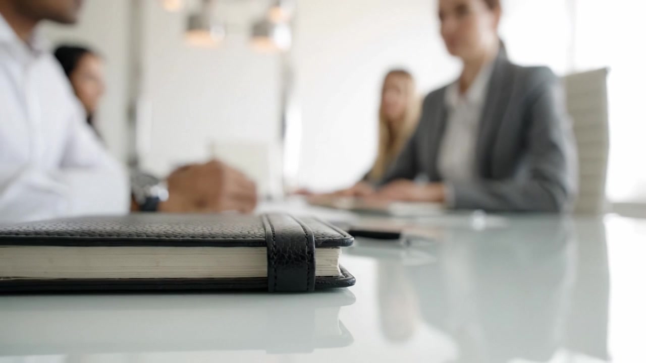 Una vista ravvicinata di un tavolo da riunione con partecipanti impegnati nella discussione e un taccuino in primo piano, che evidenzia la collaborazione professionale