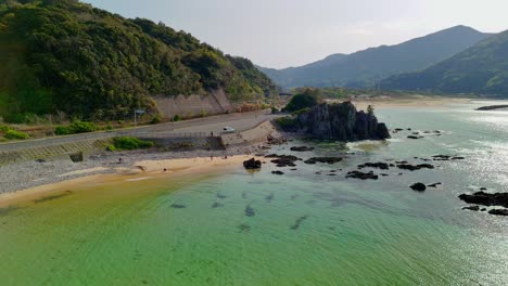 a beautiful aerial shot flies along a scenic coastal road next to a sandy beach with clear turquoise water. people can be seen enjoying the beach, with lush green mountains in the background