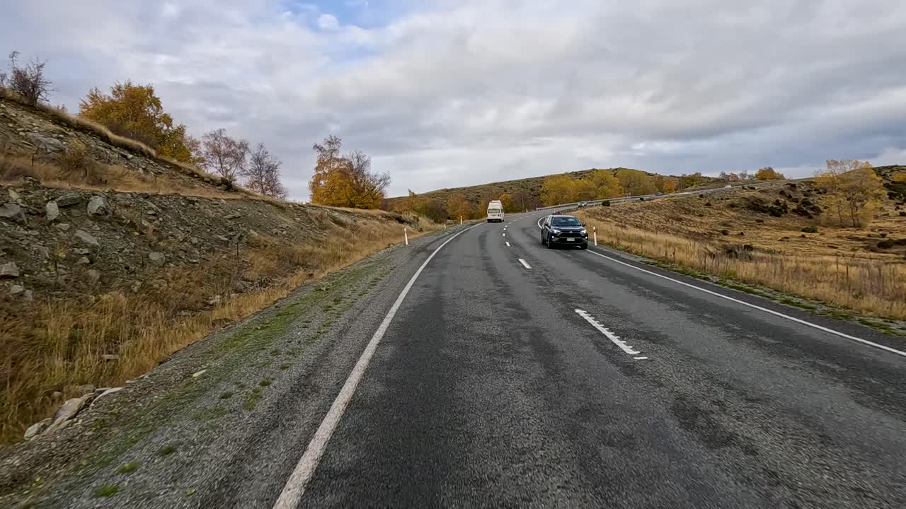 A vehicle travels a winding rural road through open farmland near Queenstown, New Zealand, under overcast skies with autumn foliage and rolling hills
