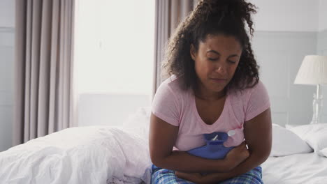 woman wearing pajamas suffering with period pain sitting on bed with hot water bottle