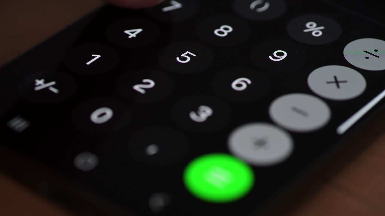 Video of a finger pressing a button on a smartphone calculator app. The bright numeric keys and green equals button stand out against the dark background, emphasizing modern digital calculation