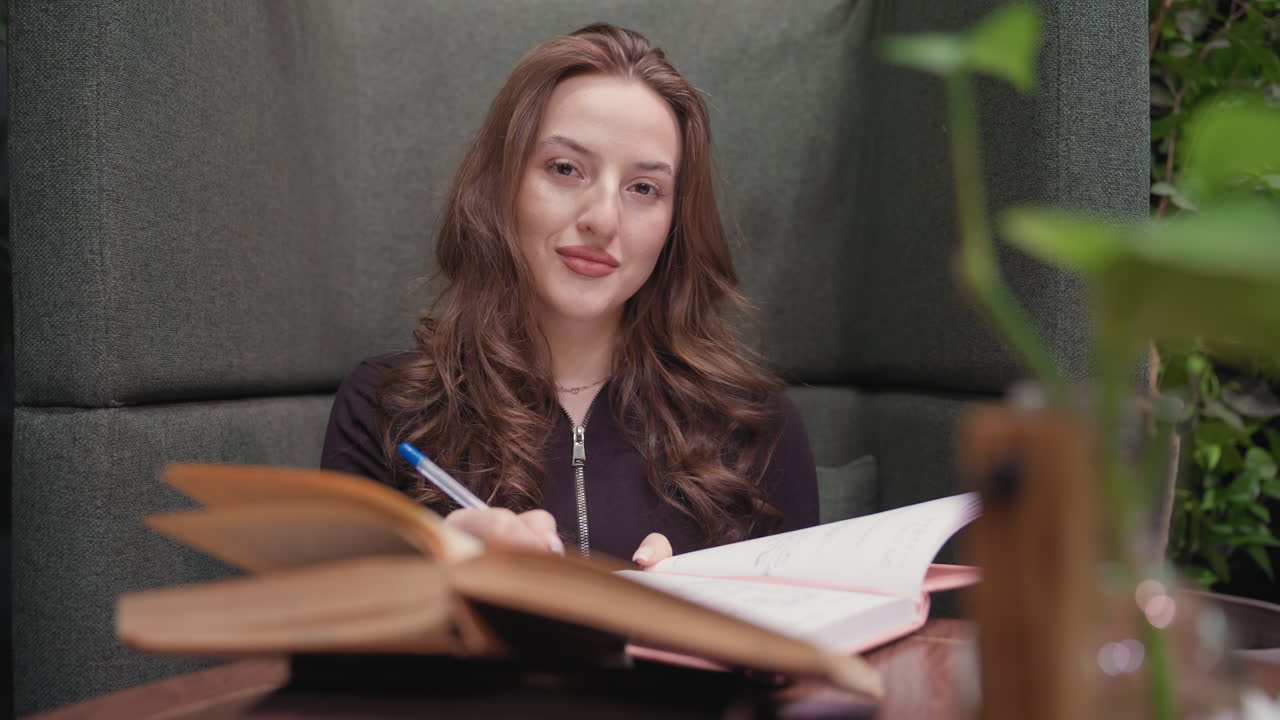 Lady with wavy brunette hair smiles gently while holding pen and looking at camera, seated in cozy green booth with open books on table, exuding calm, focused energy in relaxed indoor setting