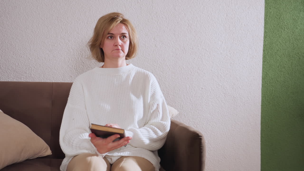 Back view of mental health professional seated while handing book to guest sitting on sofa with neutral expression, depicting calm therapeutic interaction and exchange of supportive material