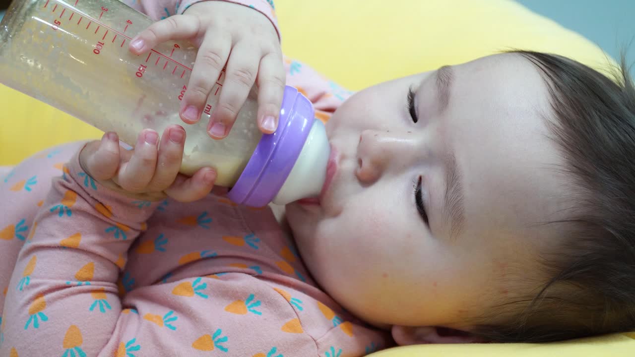 Bambina di nove mesi che tiene il biberon con latte artificiale e beve da sola sdraiata su un cuscino giallo - primo piano