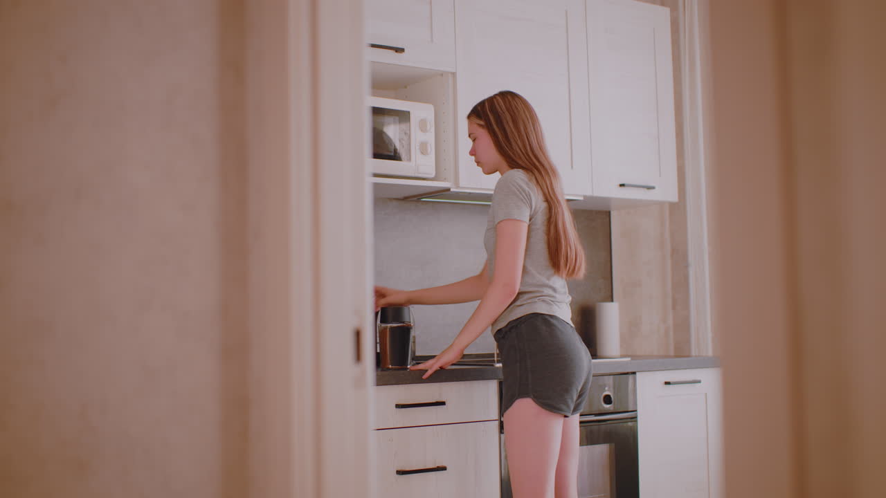 Designer prepares for work in kitchen, reaching upper cabinet to take beverage container, lowering it onto counter, planning to bring cup forward and open drawer for spoon under daylight atmosphere