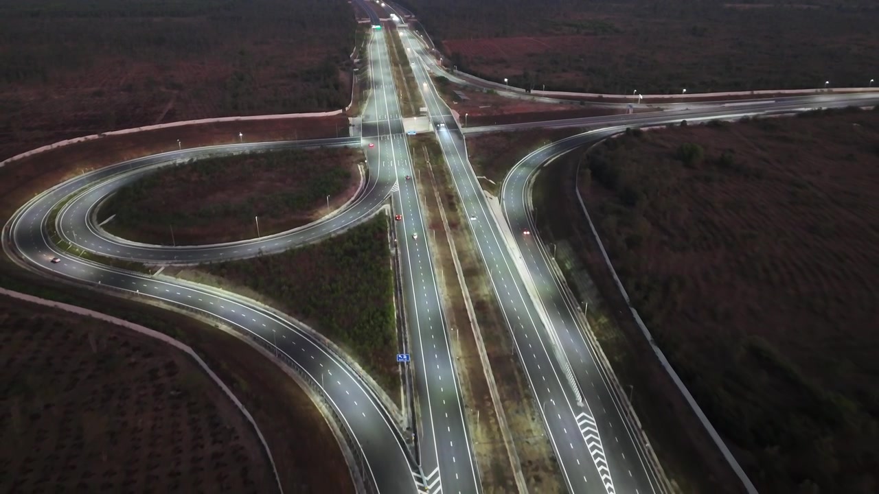 Cinematic aerial view of a modern expressway interchange illuminated at dusk, featuring sweeping curved ramps, multilane highways, and glowing roadway lights cutting through dark surrounding terrain.