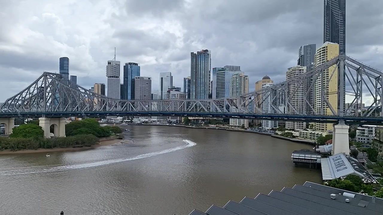 Vidvinkelbillede af Story Bridge, der strækker sig over Brisbane River med byens skyline i baggrunden. Bylandskab under overskyet himmel, med ikonisk arkitektur og flodtrafik i Brisbane.