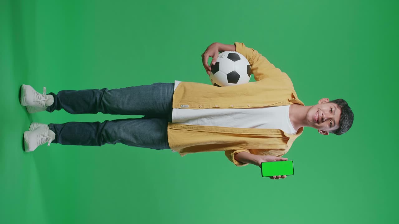 cuerpo lleno de niño asiático con una pelota que muestra un teléfono inteligente de pantalla verde y sacudiendo la cabeza mientras anima el fútbol en el fondo de la pantalla verde