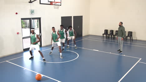 multiracial male basketball players sidestepping and warming up, coach giving instructions in gym