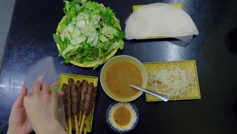 topdown shot of the hand of a female who is assembling spring roll to eat, which includes kebab, salad, noodles, and delectable gravy, on a rice paper