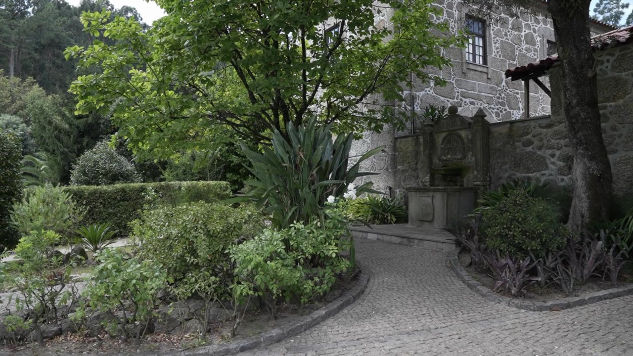 Vista del giardino di una casa in pietra con fontana, alberi, piante e un sentiero curvo in ciottoli
