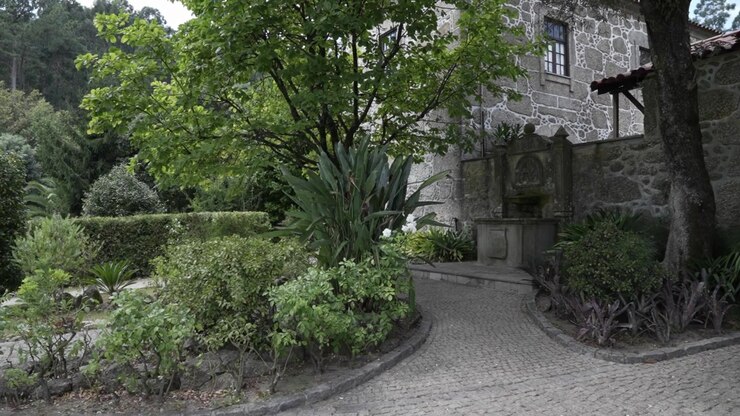 Vista del giardino di una casa in pietra con fontana, alberi, piante e un sentiero curvo in ciottoli