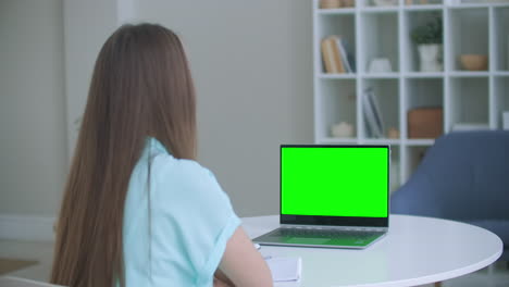 woman sits at desk in bedroom she looks at laptop green screen and talks to someone over internet video communications sometimes taking notes in notebook. close-up