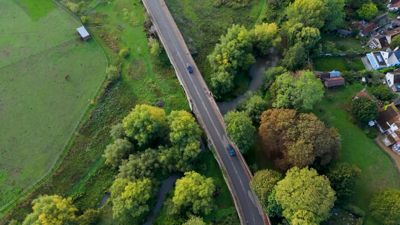 Aerial drone shot tracking an English country road. The camera moves forward, then turns left to follow the lane as cars pass below, with trees, grass, and a pond surrounding the rural landscape