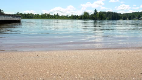 Glatte-Wellen,-Die-Langsam-Auf-Goldenes-Sandstrandufer-Rollen-Und-Abebben---Niedriger-Winkel---Dock-Und-Bäume-Im-Hintergrund