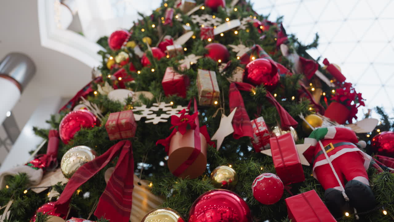 Close-up of Christmas tree with festive ornaments, golden baubles, red ribbons, decorative squirrel, and twinkling lights shining through green branches in modern shopping mall setting