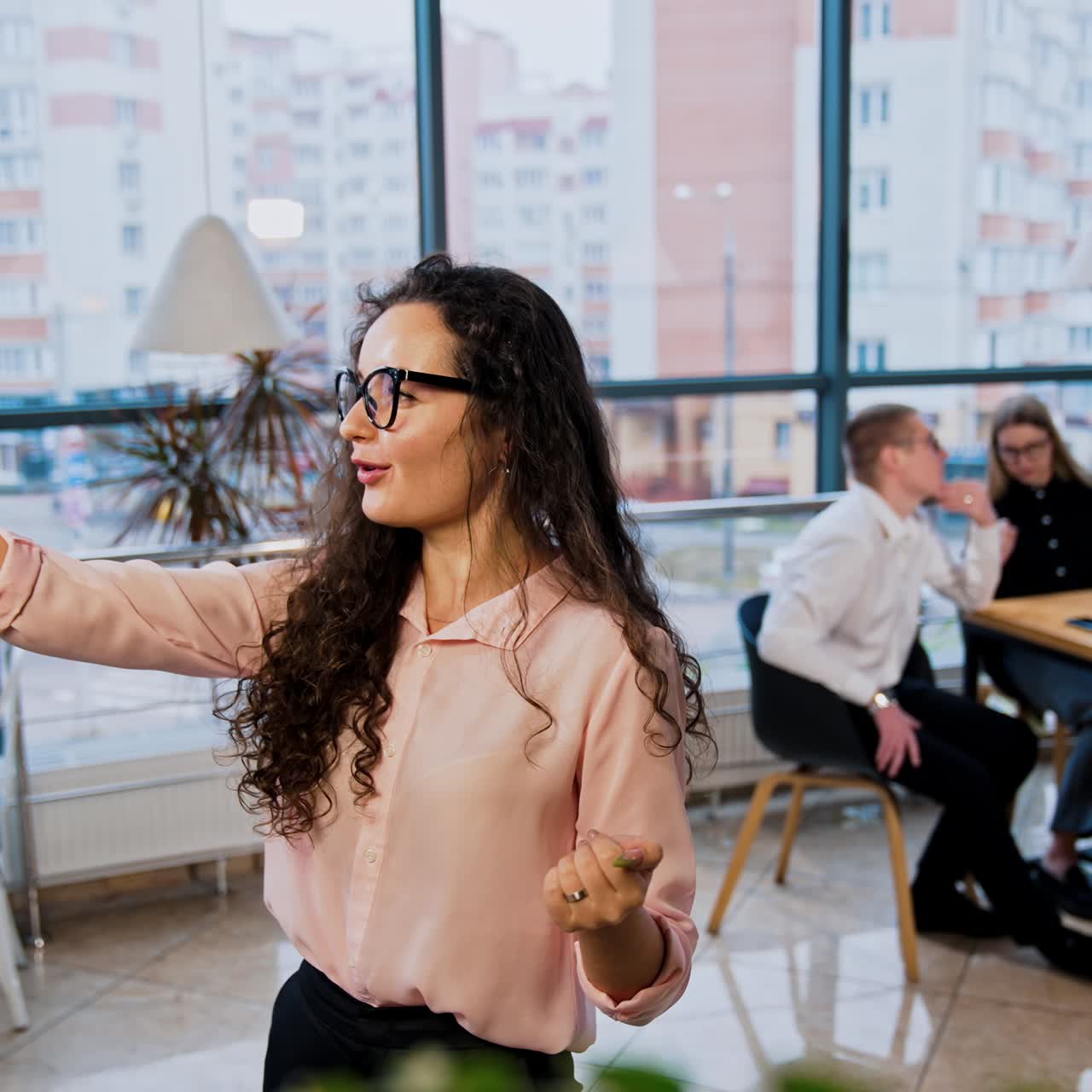 Beautiful young woman with long curly dark hair wearing glasses holding phone in hand. Lady is taking video live chat showing colleagues sitting behind at the table