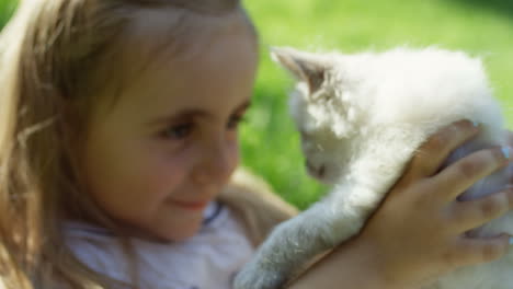 little cute girl lying on green grass and holding a little white cute kitty cat in the park