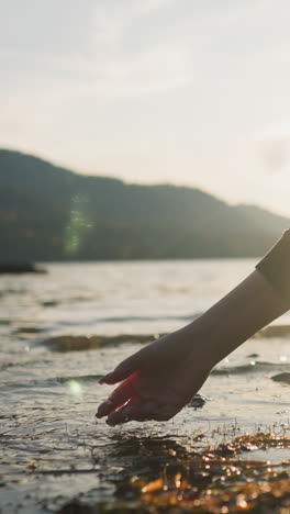lady gently strokes rippling water of lake under sunlight. woman puts hand down into warm water enjoying soft glow of sunset. connection with rhythms of nature