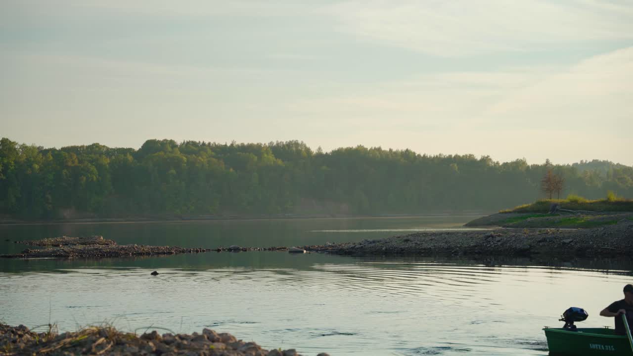 Men rowing the boat on shore. Sunset on the river with verdant forest in the background.