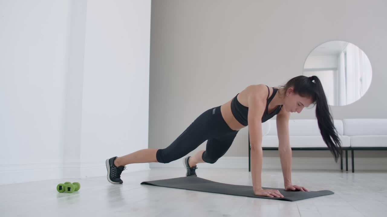 una joven morena con ropa negra y un apartamento ligero realiza el ejercicio de la tabla saltando en el resto acostado