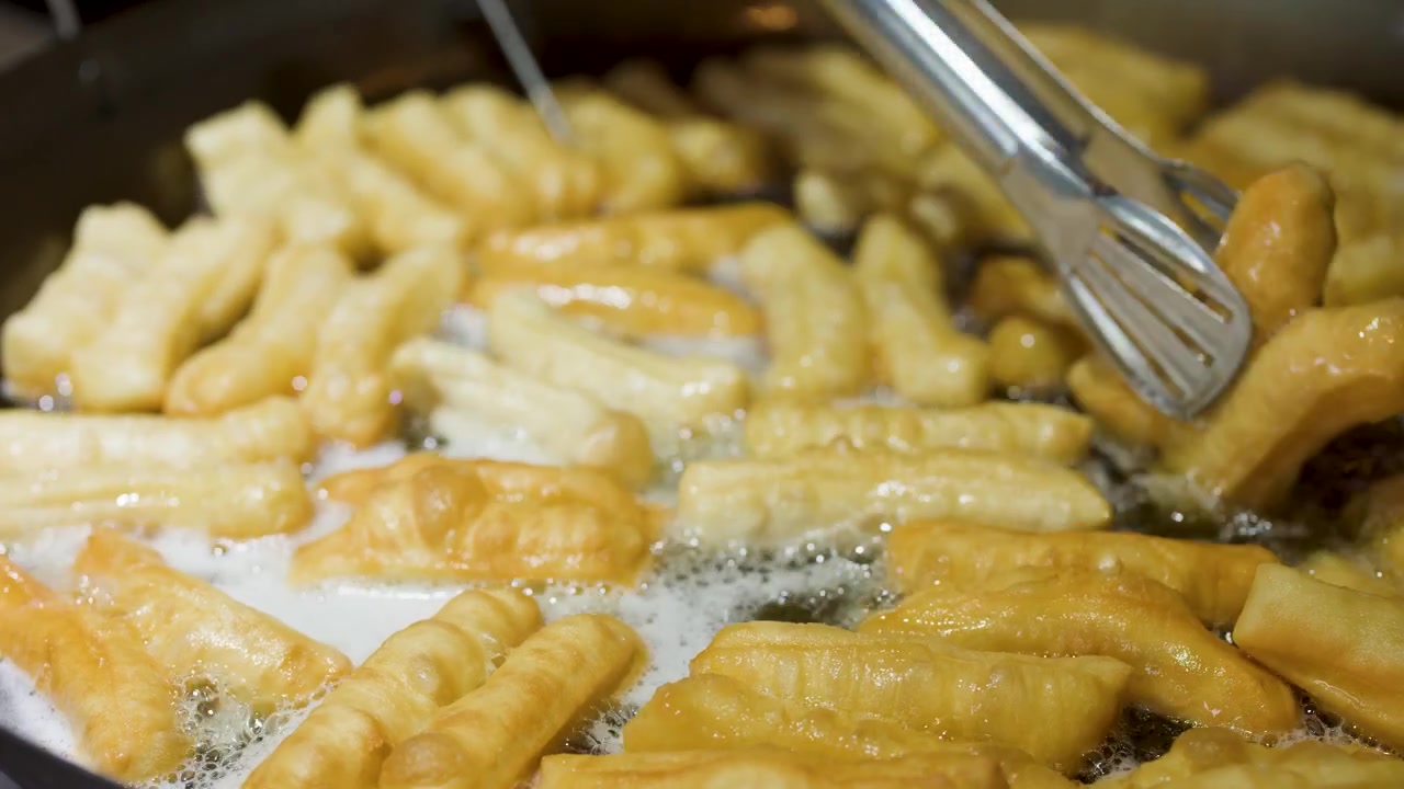 Primo piano di youtiao dorati che friggono in olio bollente, girati con pinze di metallo in un wok