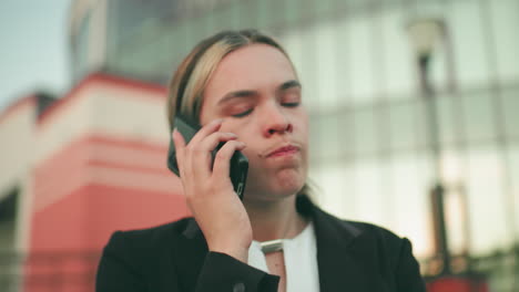 Pensive young lady in professional outfit on phone call appearing tired and thoughtful, with slightly blurred modern building and two lampstands in background