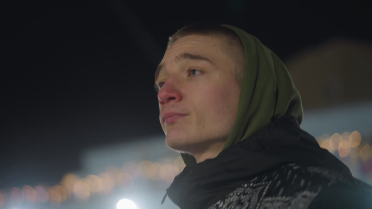 Moody man in green hoodie and patterned winter jacket walking outdoors at night, looking focused with cold breath visible in frosty air, warm bokeh lights glowing softly in background
