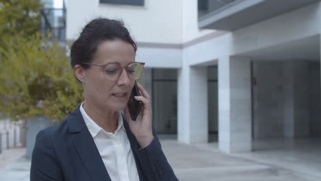 serious businesswoman in office suit talking on mobile phone while standing outside near office building