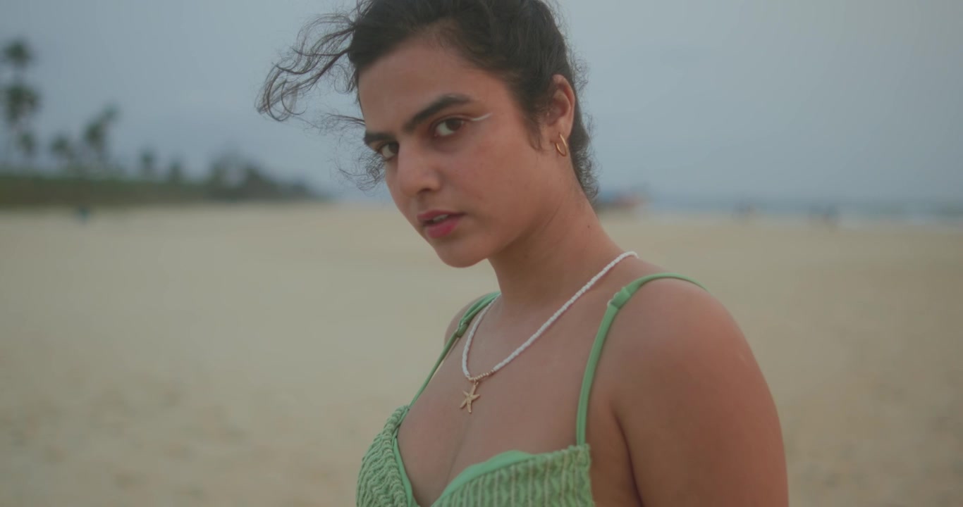 Giovane donna in piedi sulla spiaggia che guarda di lato in una luce naturale soffusa. Ritratto lifestyle cinematografico con un'atmosfera calma, ideale per viaggi, moda e narrazione emotiva.