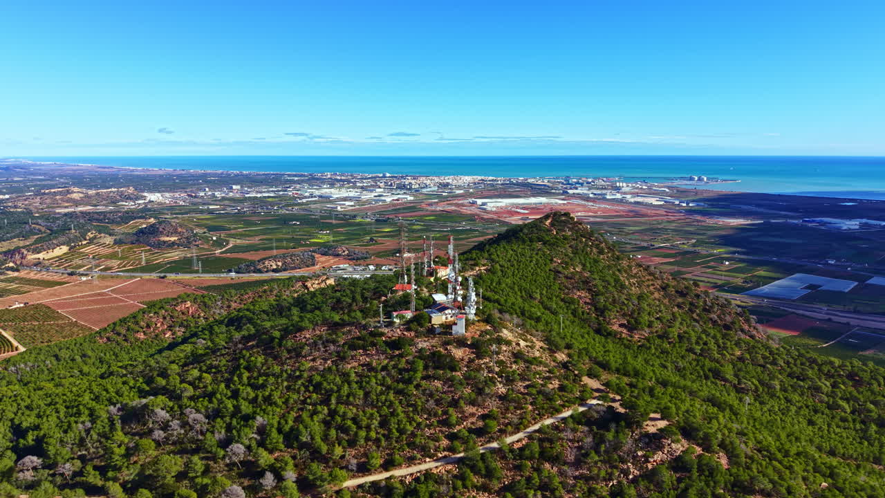 Veduta di tralicci per le telecomunicazioni su una collina che domina una città costiera con campi e fattorie sullo sfondo, di giorno.