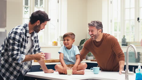 Familia-Del-Mismo-Sexo-Con-Dos-Papás-En-La-Cocina-Y-Un-Hijo-Sentado-En-El-Mostrador.