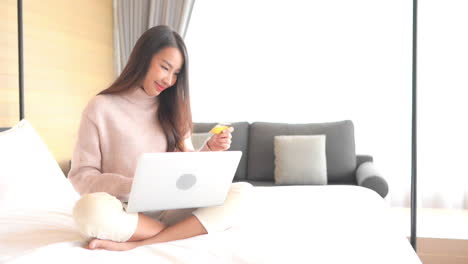 asian woman with a beautiful smile is using her credit card to make an online payment using a laptop computer, money transfer, female sitting cross-legged on the bed and put in credit card info