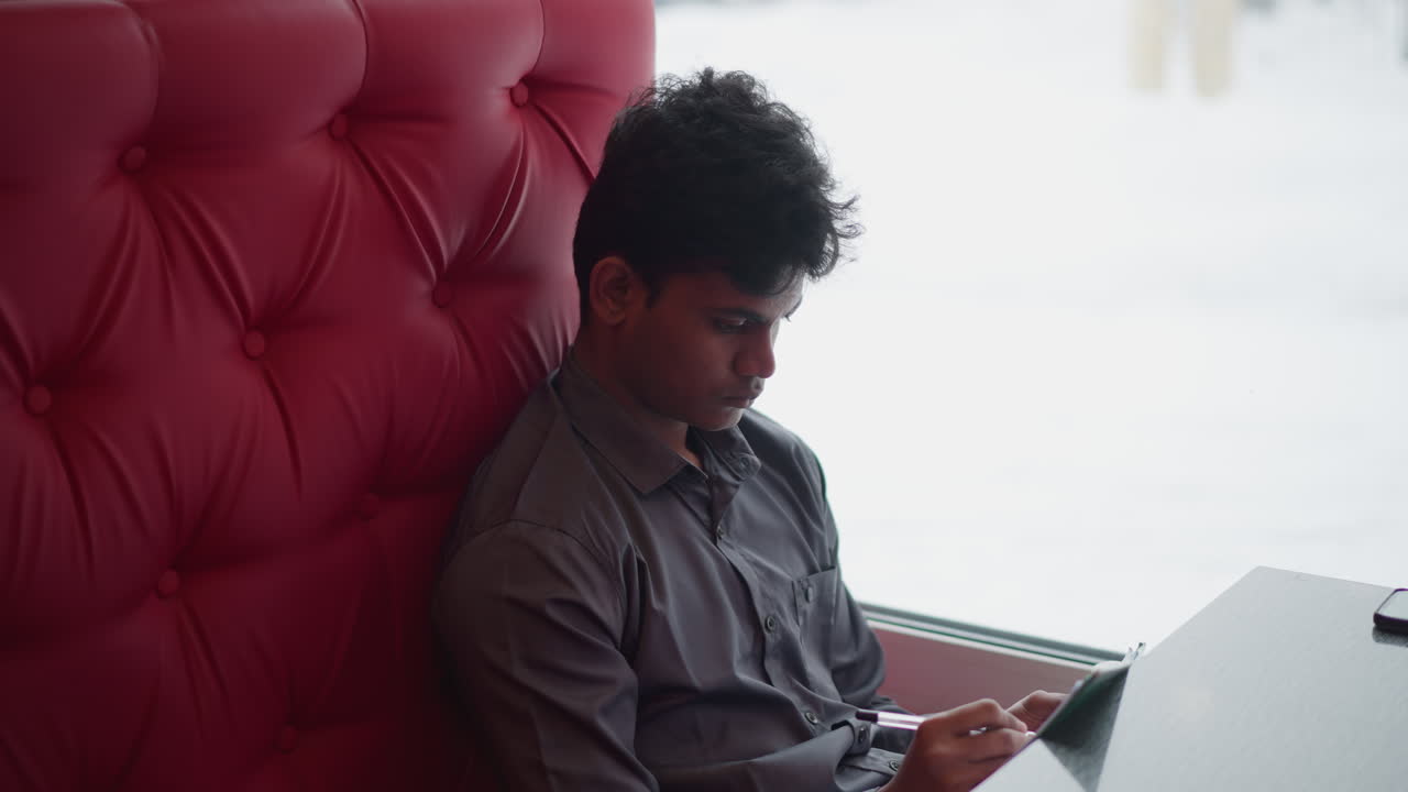 Student sitting on red cushioned bench by window concentrating on notebook, holding pen while writing or drawing, winter scene visible outside, peaceful atmosphere perfect for creativity or academic focus