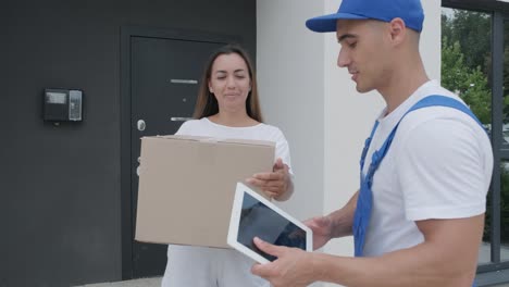 beautiful woman meets a delivery man who gives her a parcel box beside her home, she signs electronic signature on tablet. concept of courier, home delivery, online shopping