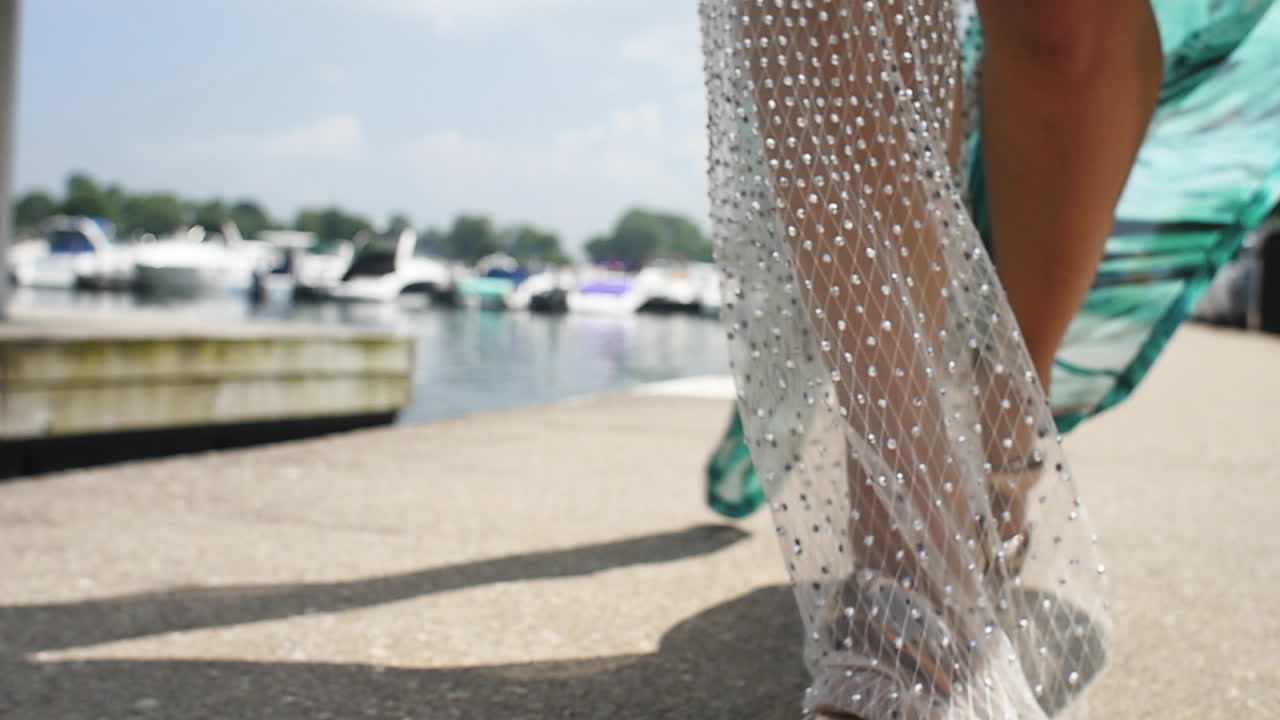 여름 햇살 아래 보트 선착장을 따라 걷는 동안 고급스러운 여성이 황금 굽 신발을 신습니다.