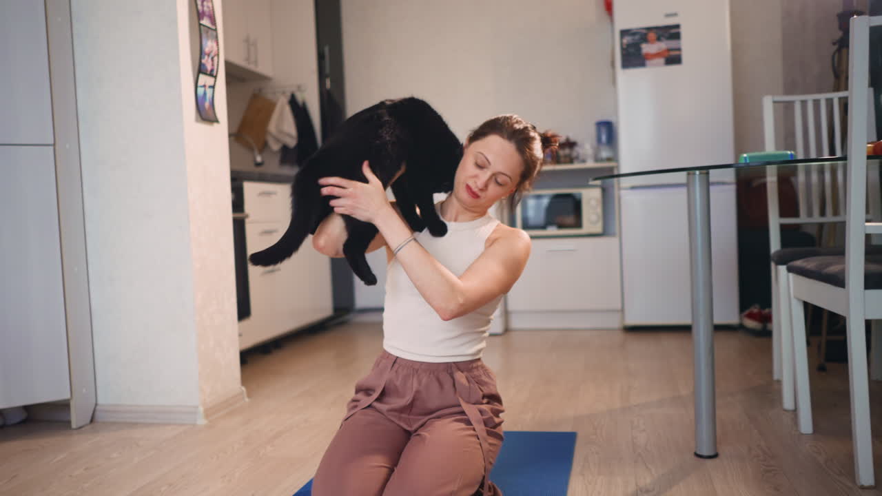 Caucasian Instructor Coaxing Black Cat Onto Yoga Mat, Kneeling On Blue Mat In Bright WoodenFloor Apartment, Playful Lift And Gentle Guidance, Tutorial Vibe For PetFriendly Practice