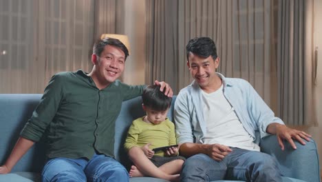 asian gay fathers and their cute son relaxing on couch and using mobile phone and warmly smiling to camera