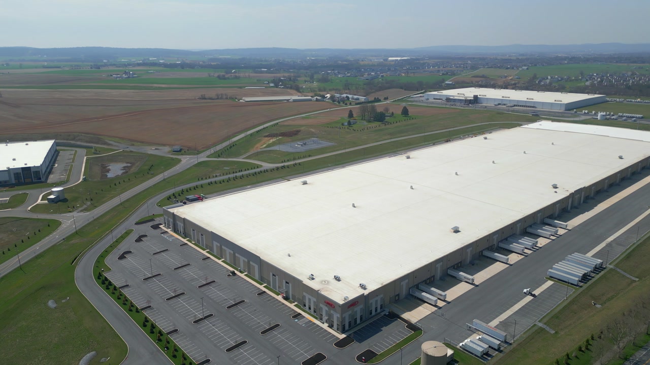 Cinematic drone shot of a large modern industrial warehouse and commercial building. High angle view of the facility surrounded by green fields and trees under an overcast sky.