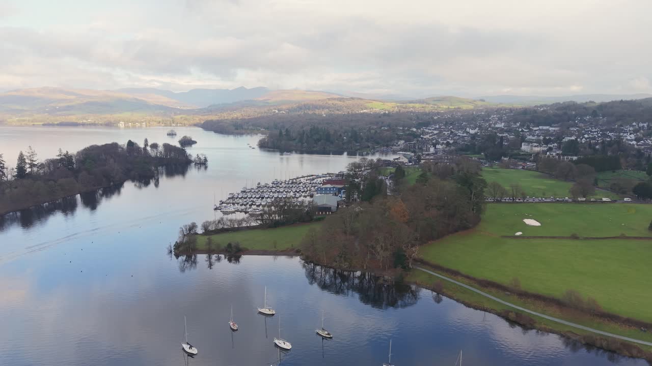 Geser ke kiri melintasi Danau Windermere dengan daratan di sebelah kanan dan perairan terbuka di bawahnya. Drone melintas ke arah marina kecil saat cahaya matahari menembus awan, menerangi puncak gunung yang jauh