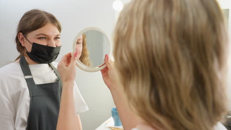 beautician wearing black face mask hands mirror to blonde client for eyebrow check. client reflection appears in mirror as both stand in well-lit beauty salon with white background and soft lighting